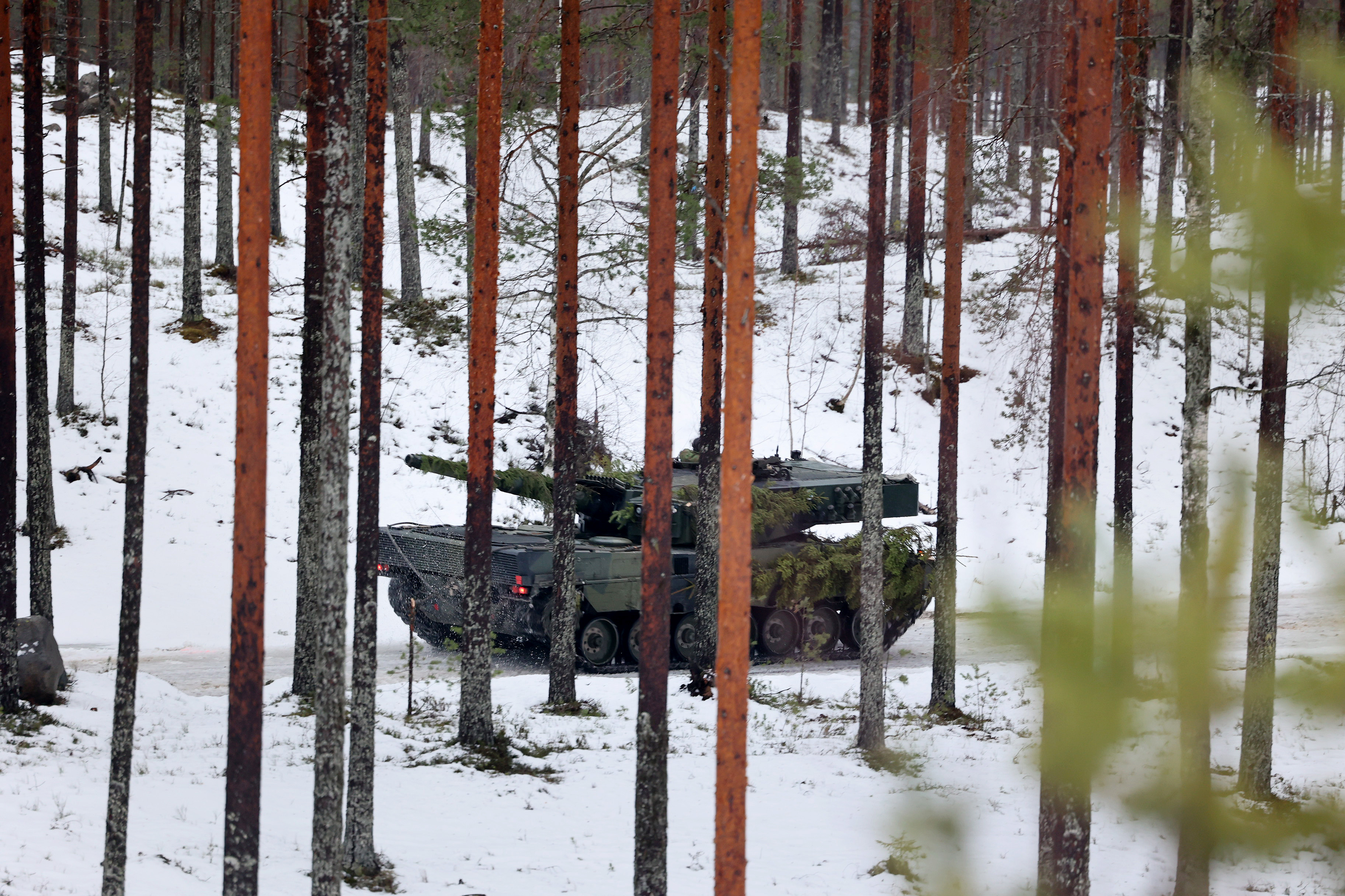 A military vehicle camouflaged with foliage traverses a snowy forest, surrounded by tall pine trees, creating a still, cold, and secretive atmosphere.