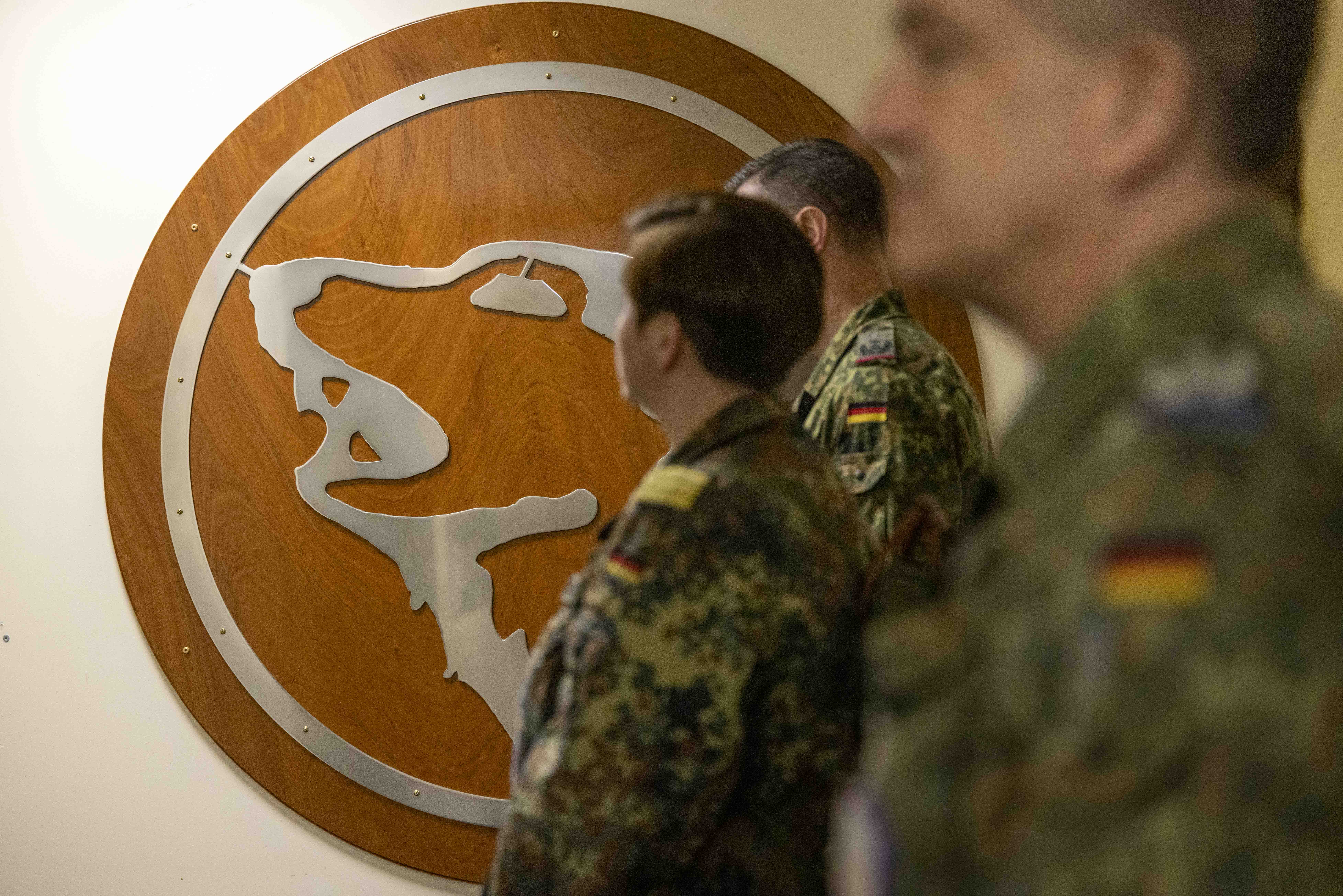 Soldiers in camo uniforms stand in front of a wooden wall displaying a silver bear insignia. The atmosphere is formal and focused.