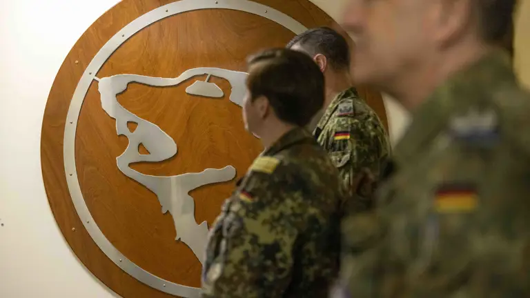 Soldiers in camo uniforms stand in front of a wooden wall displaying a silver bear insignia. The atmosphere is formal and focused.