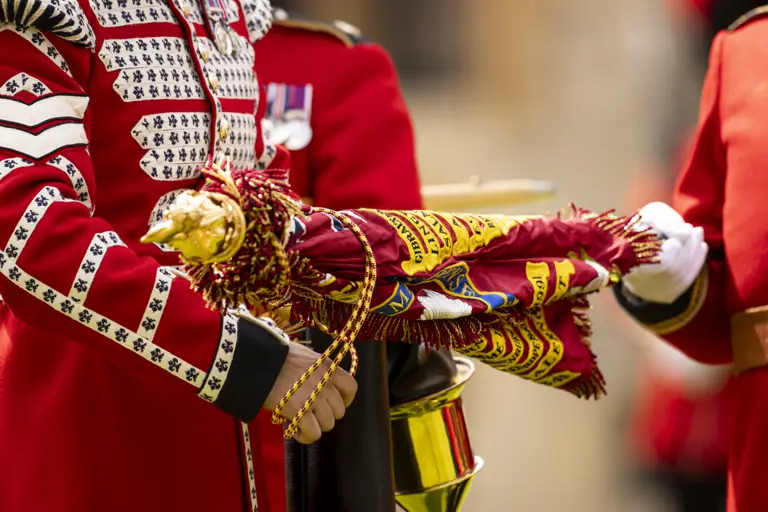 Close-up of a soldier in a red ceremonial uniform holding a decorated regimental flag with gold fringe. 