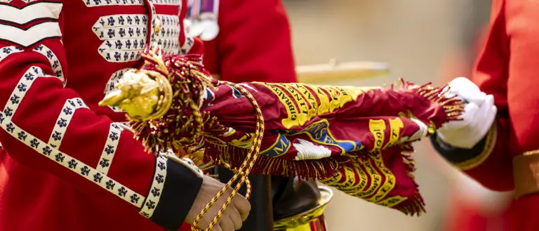 Close-up of a soldier in a red ceremonial uniform holding a decorated regimental flag with gold fringe.