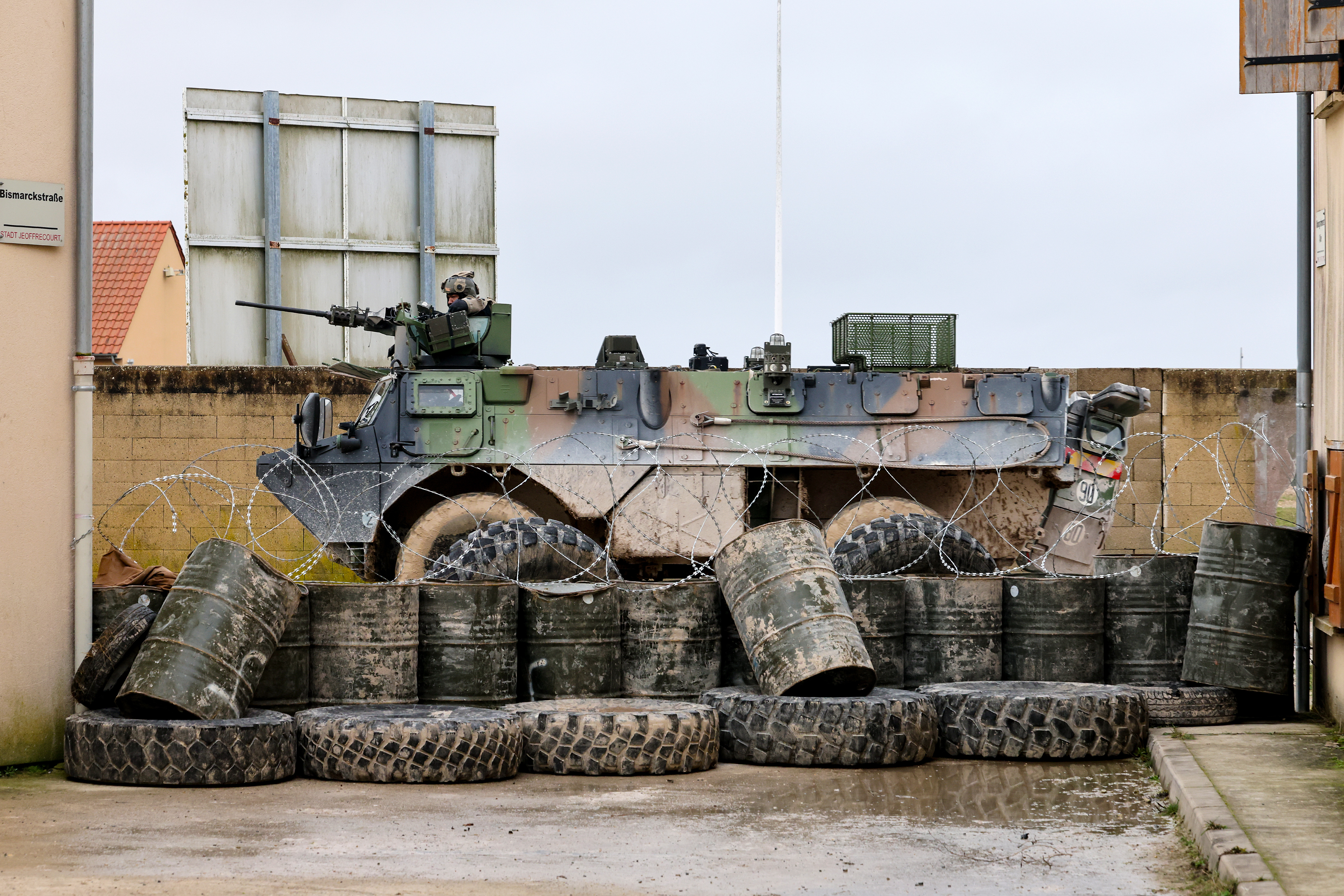Camouflaged military armored vehicle parked behind a barricade of tires and metal barrels topped with barbed wire.