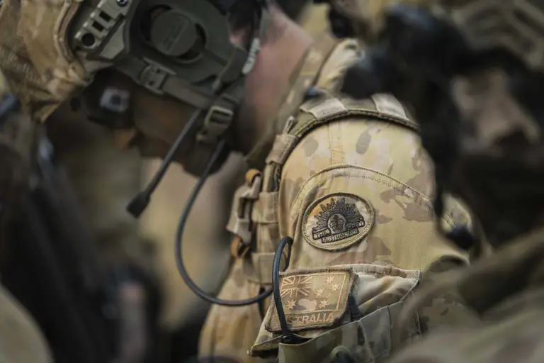 A soldier wearing camouflage gear and a communication headset, focusing intently. His uniform includes visible Australian military patches.