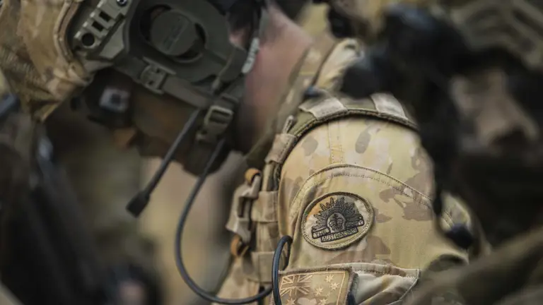 A soldier wearing camouflage gear and a communication headset, focusing intently. His uniform includes visible Australian military patches.
