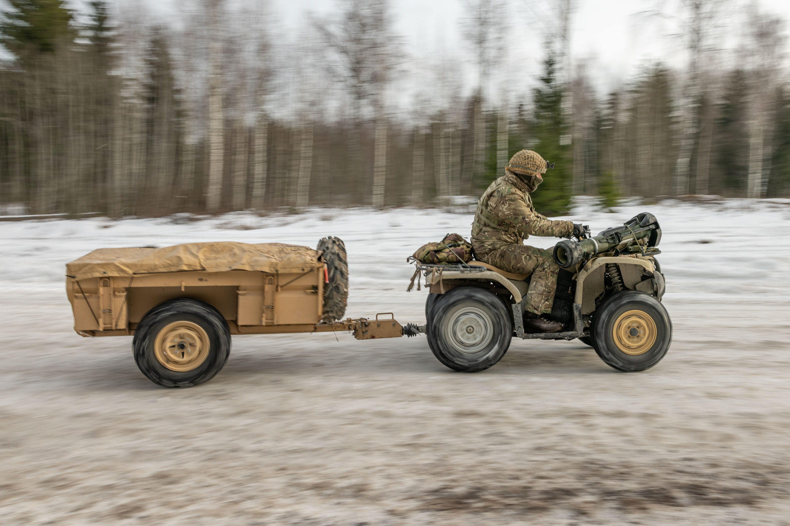 A person in camouflage clothing drives an ATV towing a covered trailer through a snowy, forested landscape. 
