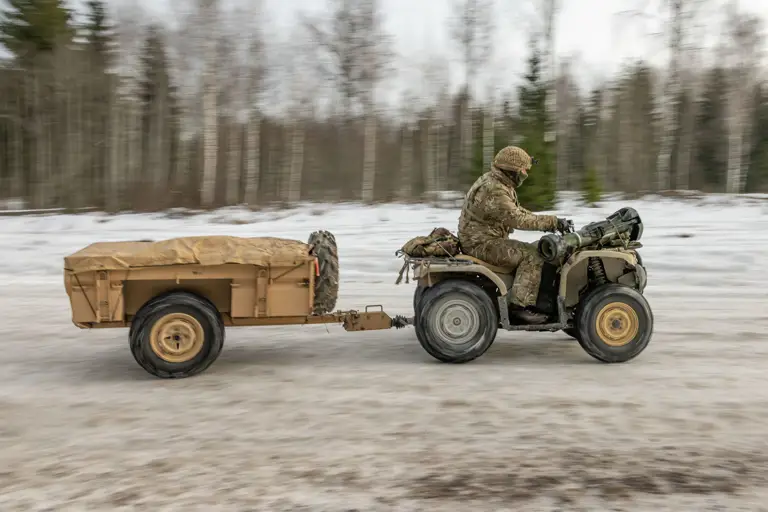A person in camouflage clothing drives an ATV towing a covered trailer through a snowy, forested landscape.