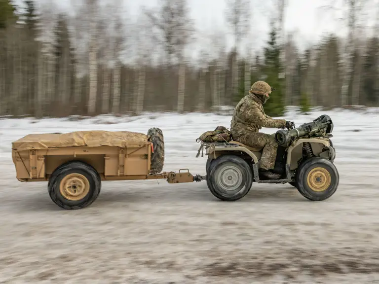 A person in camouflage clothing drives an ATV towing a covered trailer through a snowy, forested landscape.