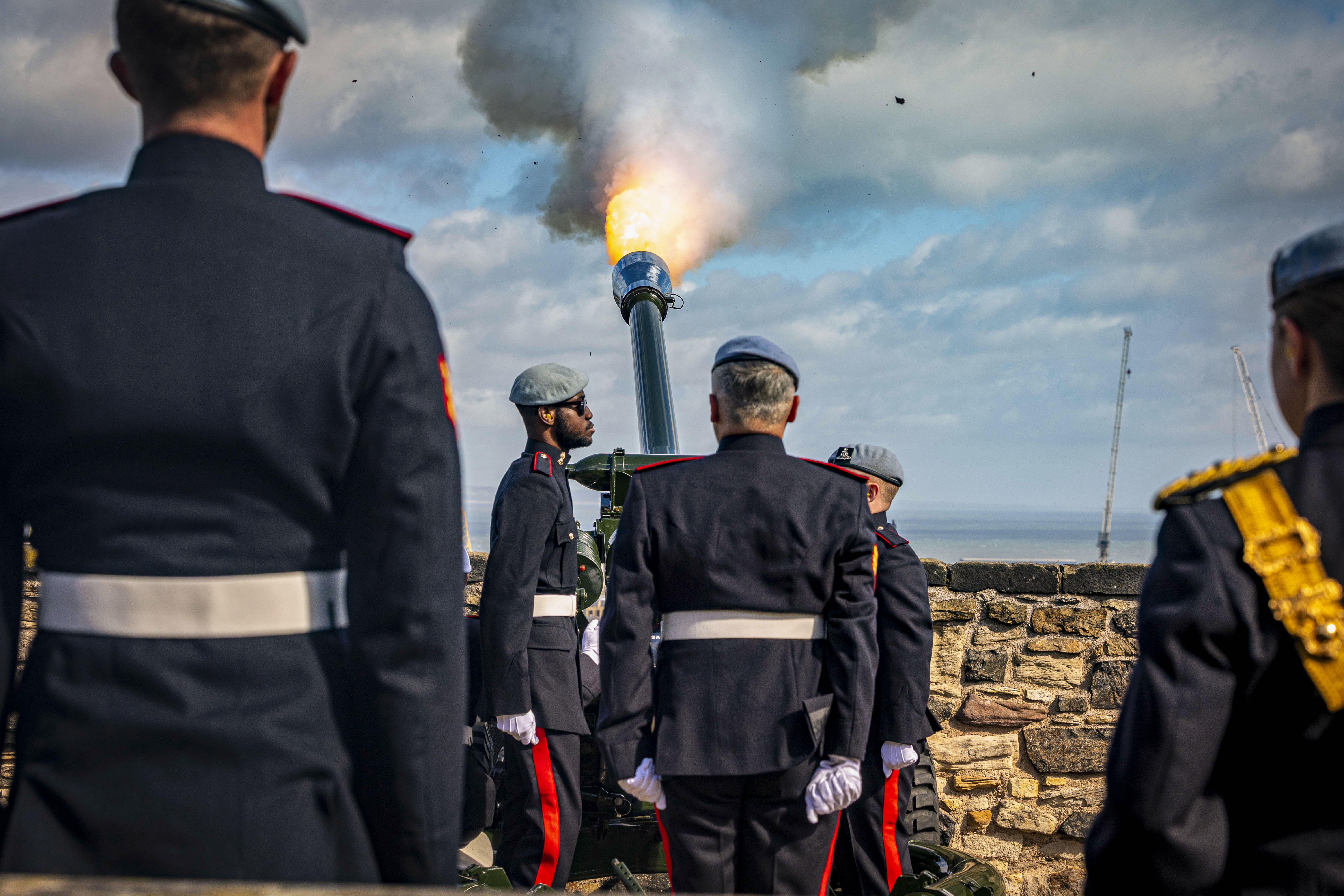 A group of uniformed individuals stand around a cannon firing at Stirling Castle. The cannon emits smoke and fire against a blue sky. The scene is near a stone wall with a view of the sea in the background.