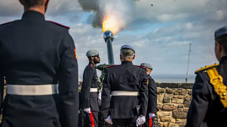 A group of uniformed individuals stand around a cannon firing at Stirling Castle. The cannon emits smoke and fire against a blue sky. The scene is near a stone wall with a view of the sea in the background.