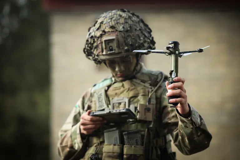 A man in uniform holds a drone and looks down at a portable screen.