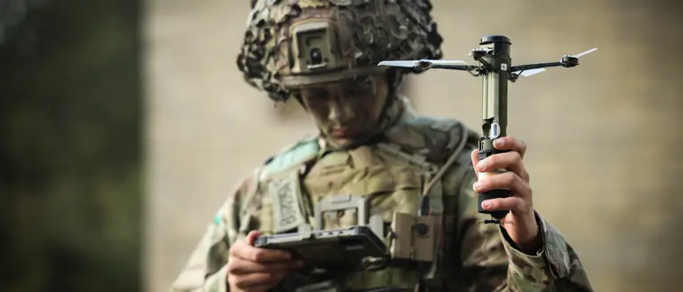 A man in uniform holds a drone and looks down at a portable screen.
