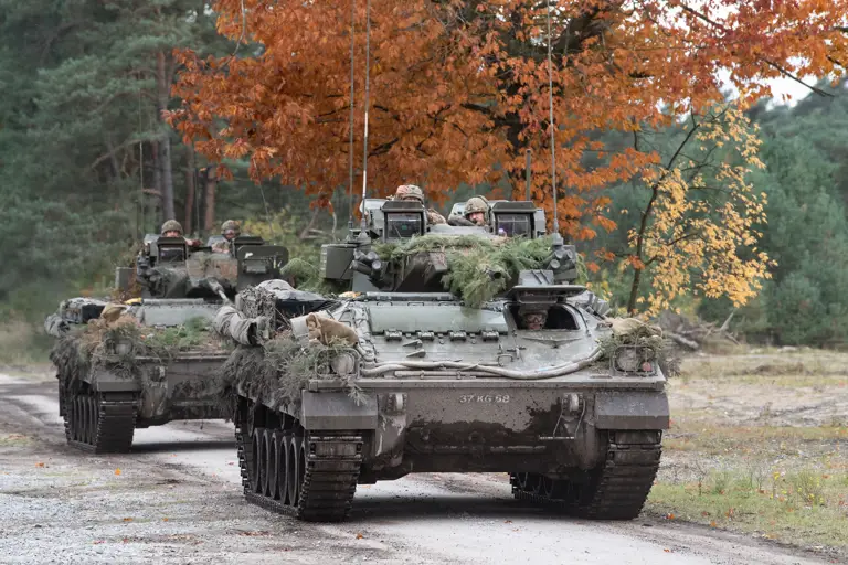 Two green armoured vehicles drive along a concrete road.
