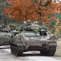 Two green armoured vehicles drive along a concrete road.