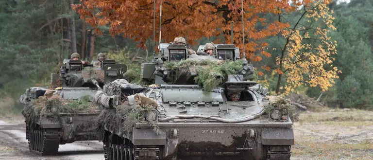 Two green armoured vehicles drive along a concrete road.
