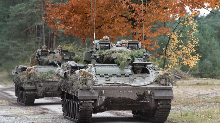Two green armoured vehicles drive along a concrete road.