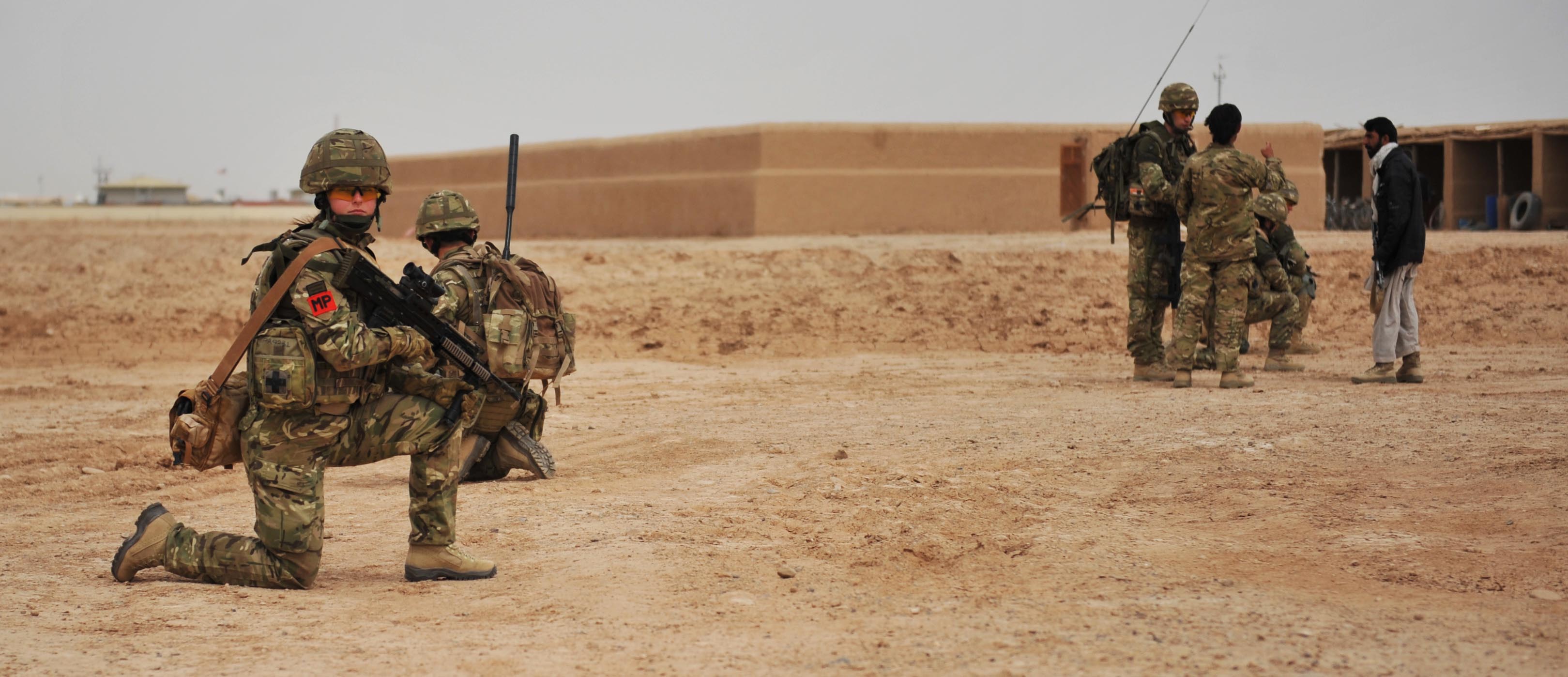 A group of military personnel in camouflage uniforms are in a desert landscape. Some are kneeling with their weapons, while others are standing and talking to a person in civilian clothing. An adobe building is visible in the background.