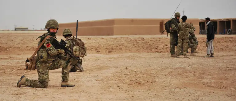 A group of military personnel in camouflage uniforms are in a desert landscape. Some are kneeling with their weapons, while others are standing and talking to a person in civilian clothing. An adobe building is visible in the background.