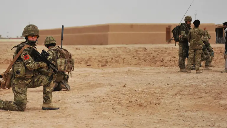 A group of military personnel in camouflage uniforms are in a desert landscape. Some are kneeling with their weapons, while others are standing and talking to a person in civilian clothing. An adobe building is visible in the background.