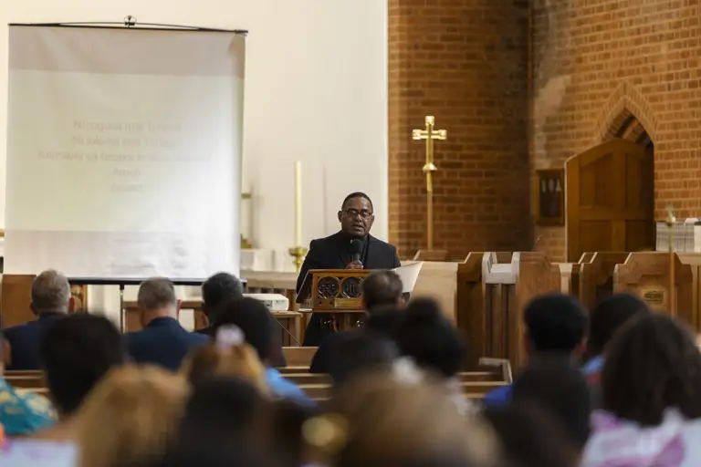 A speaker stands at a wooden lectern addressing a seated congregation inside a brick-walled church.