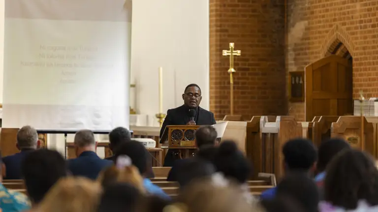 A speaker stands at a wooden lectern addressing a seated congregation inside a brick-walled church.