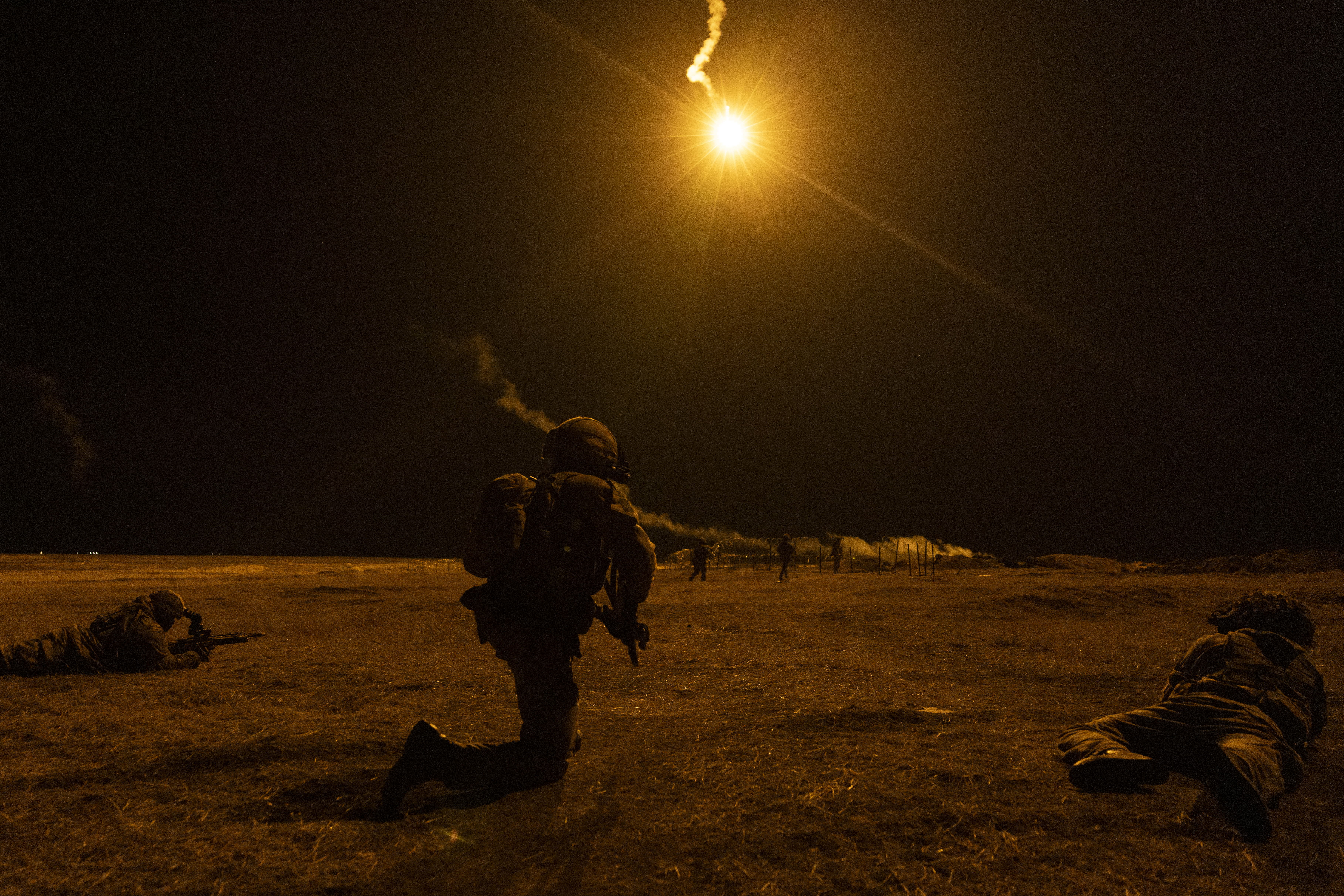 Soldiers on exercise seem during a night firing exercise, one soldier is seen on the ground, another on their knee with flares in the sky.