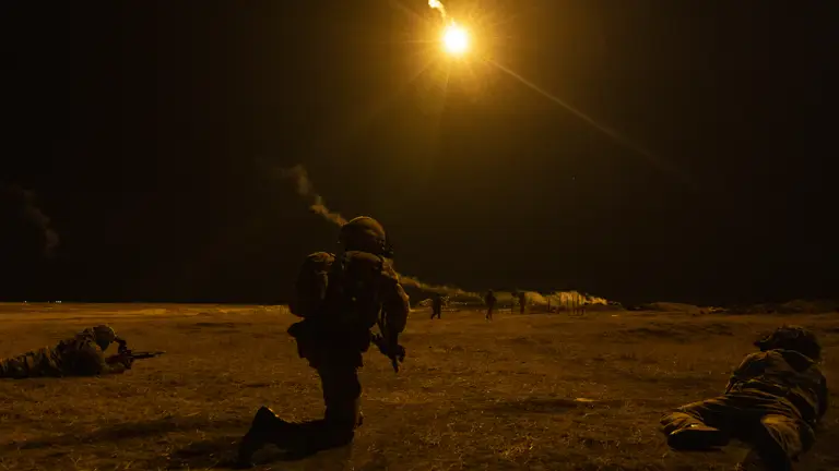Soldiers on exercise seem during a night firing exercise, one soldier is seen on the ground, another on their knee with flares in the sky.