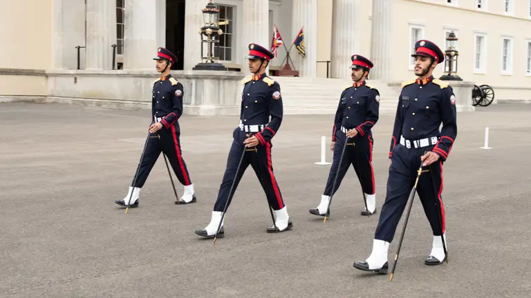 Four Qatar Defence Force soldiers wearing dress uniforms are marching across a parade square using pace sticks. Behind them are the steps of the Royal Military Academy Sandhurst.