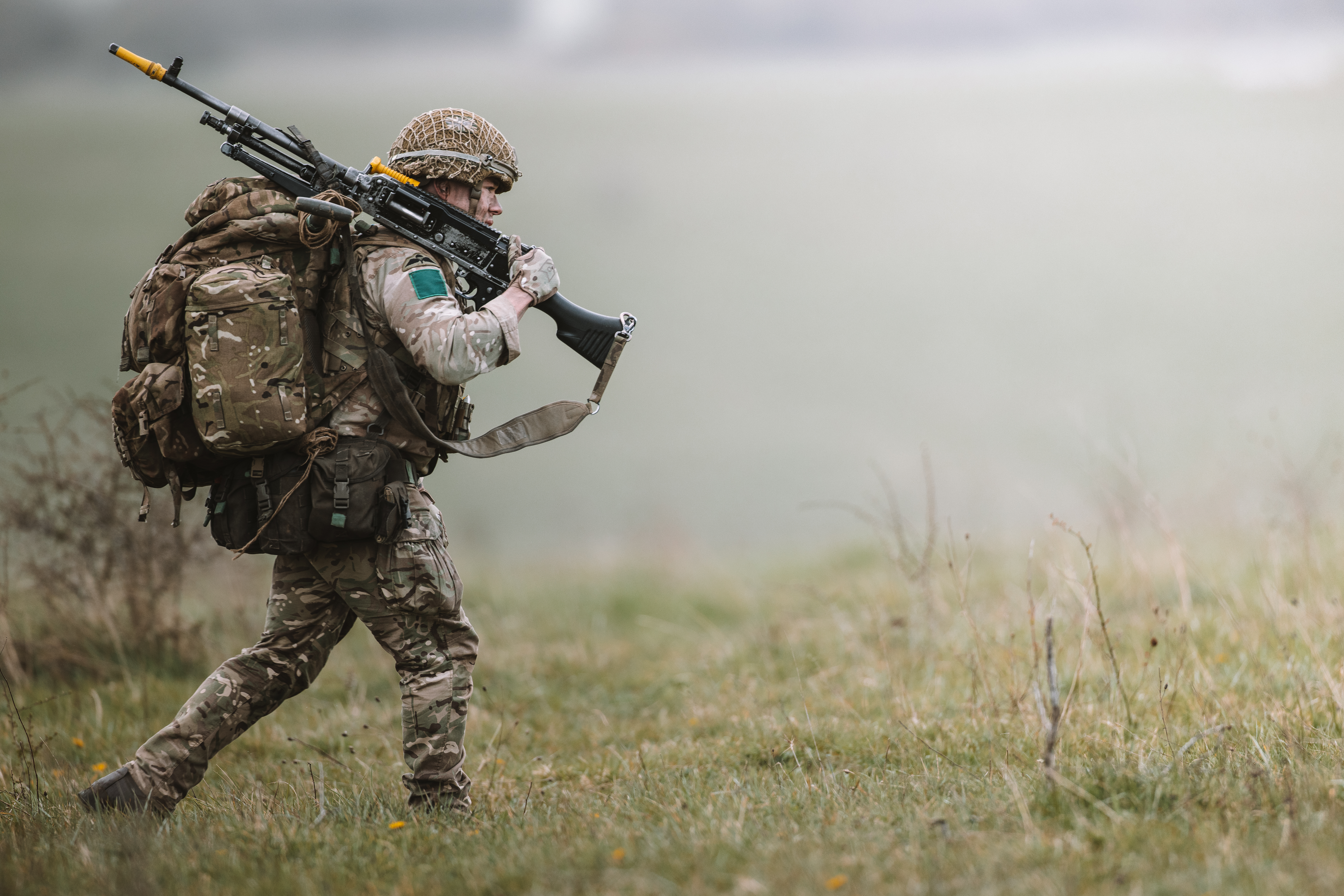 A soldier in camouflage gear, carrying a large backpack and a rifle, moves across a grassy field. 