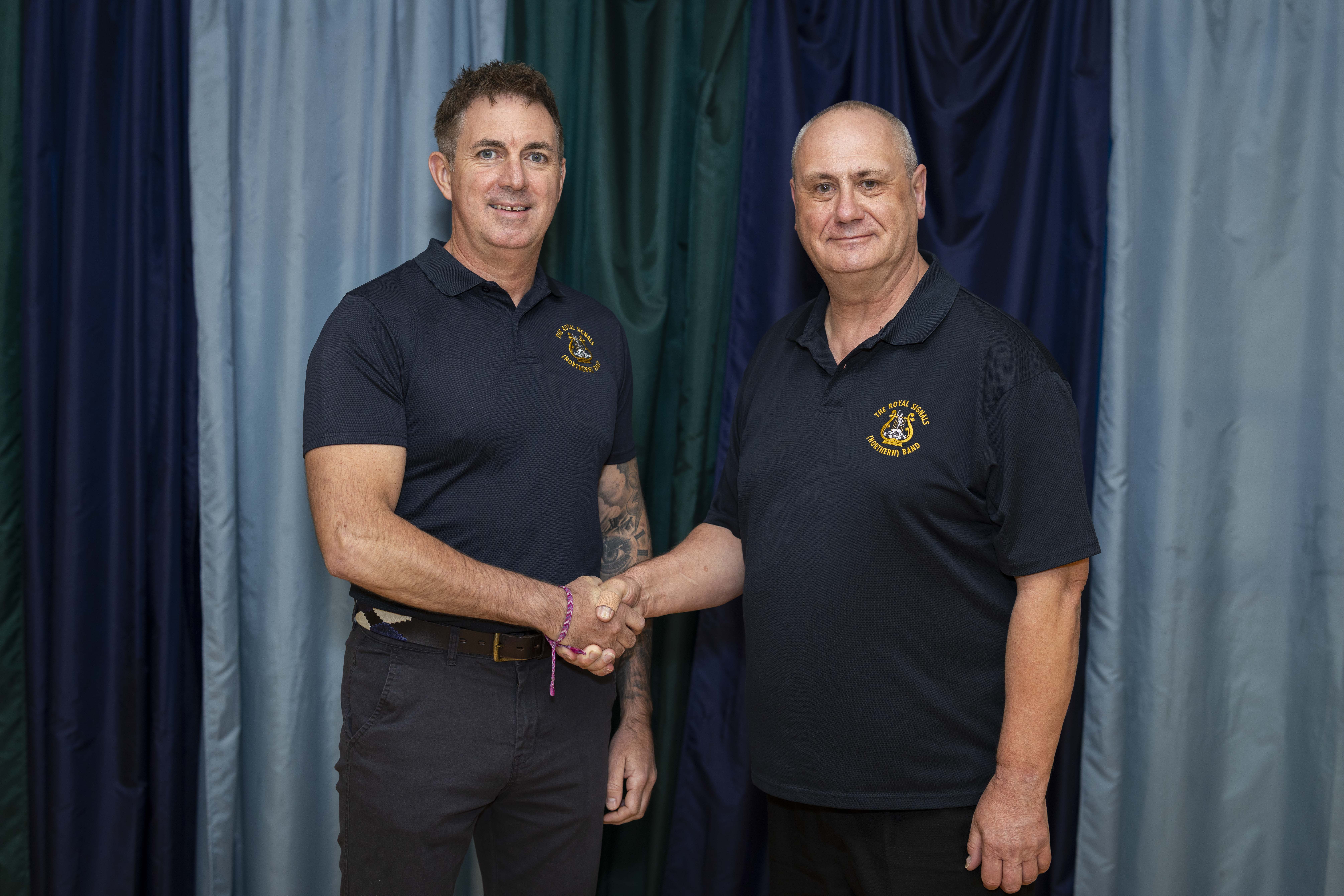 Two people are shaking hands in front of a curtain backdrop. Both are wearing dark polo shirts with embroidered logos.