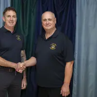 Two people are shaking hands in front of a curtain backdrop. Both are wearing dark polo shirts with embroidered logos.