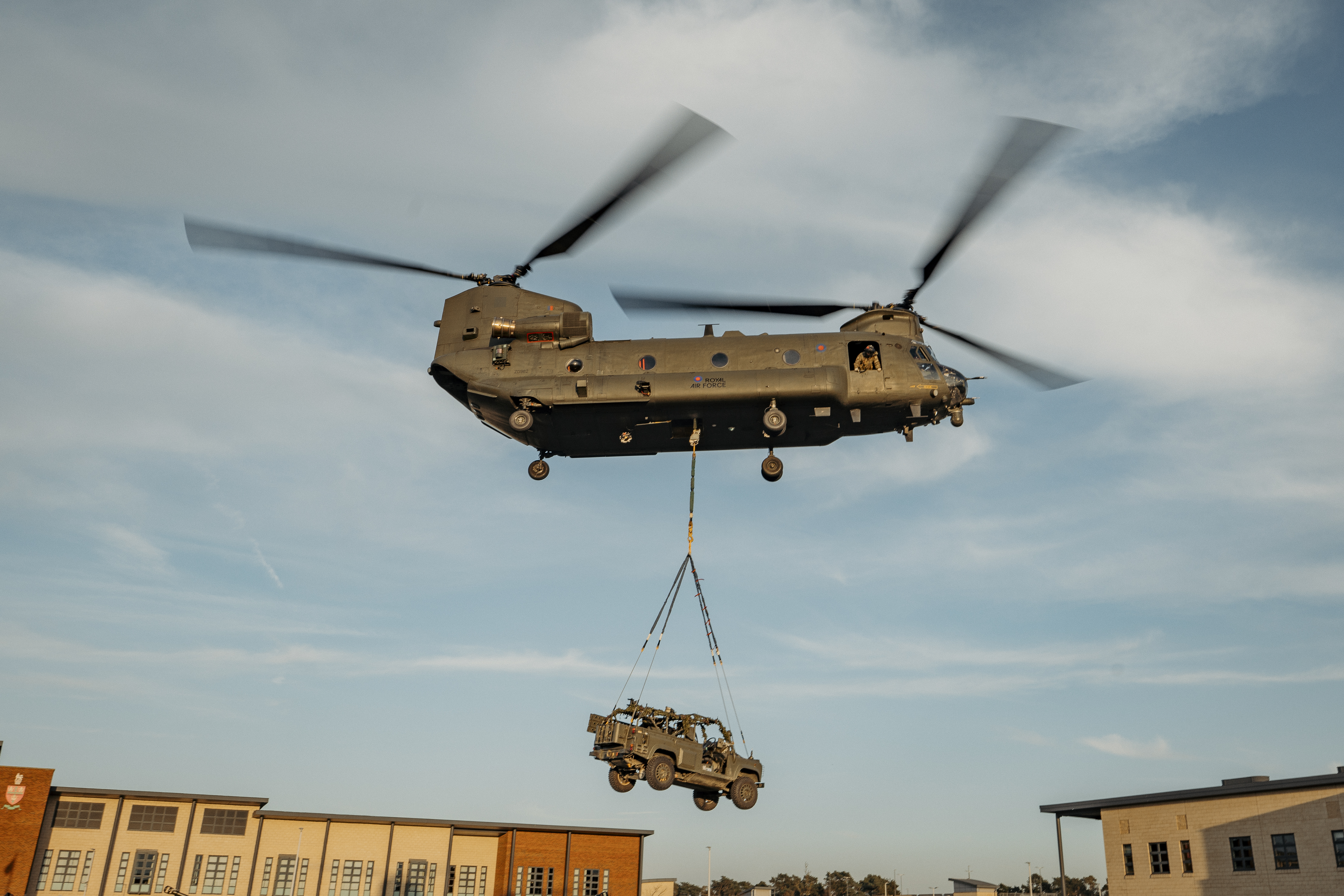 Chinook is seen flying over army barracks transporting an army vehicle attached to it's underslung. 