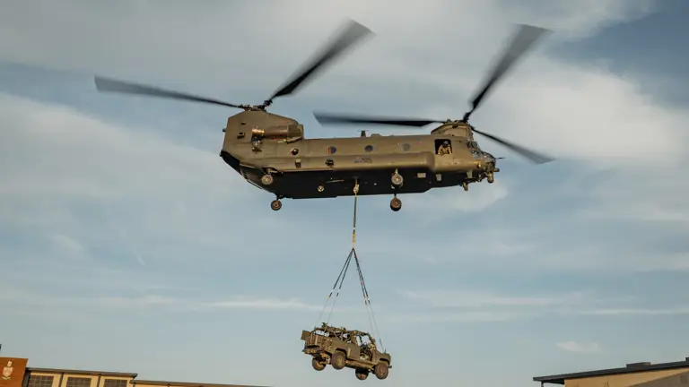 Chinook is seen flying over army barracks transporting an army vehicle attached to it's underslung.