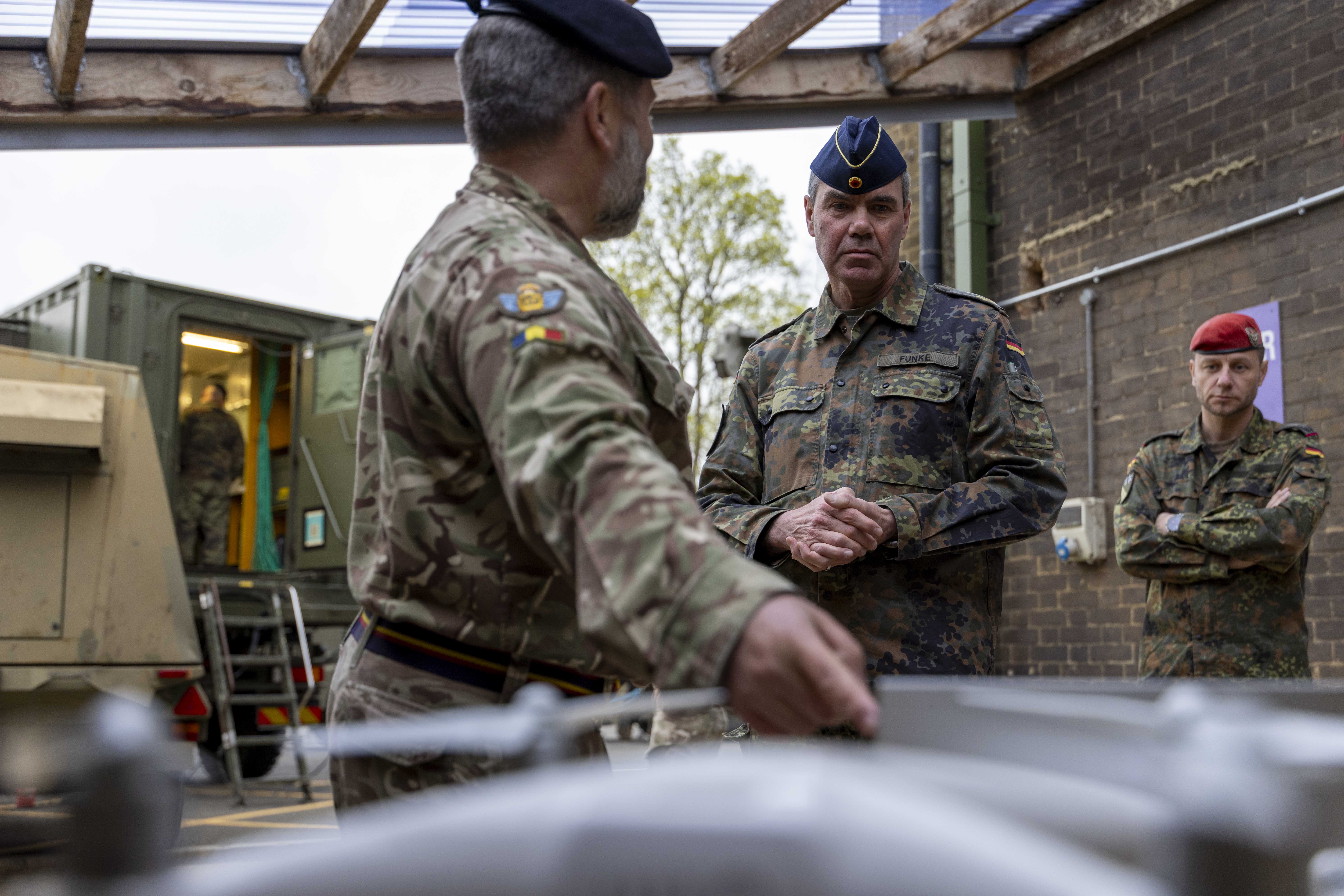 Military officers in camo uniforms and berets converse in an outdoor setting with portable military equipment. The mood is serious and focused.