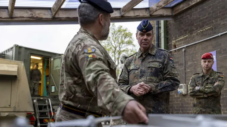 Military officers in camo uniforms and berets converse in an outdoor setting with portable military equipment. The mood is serious and focused.