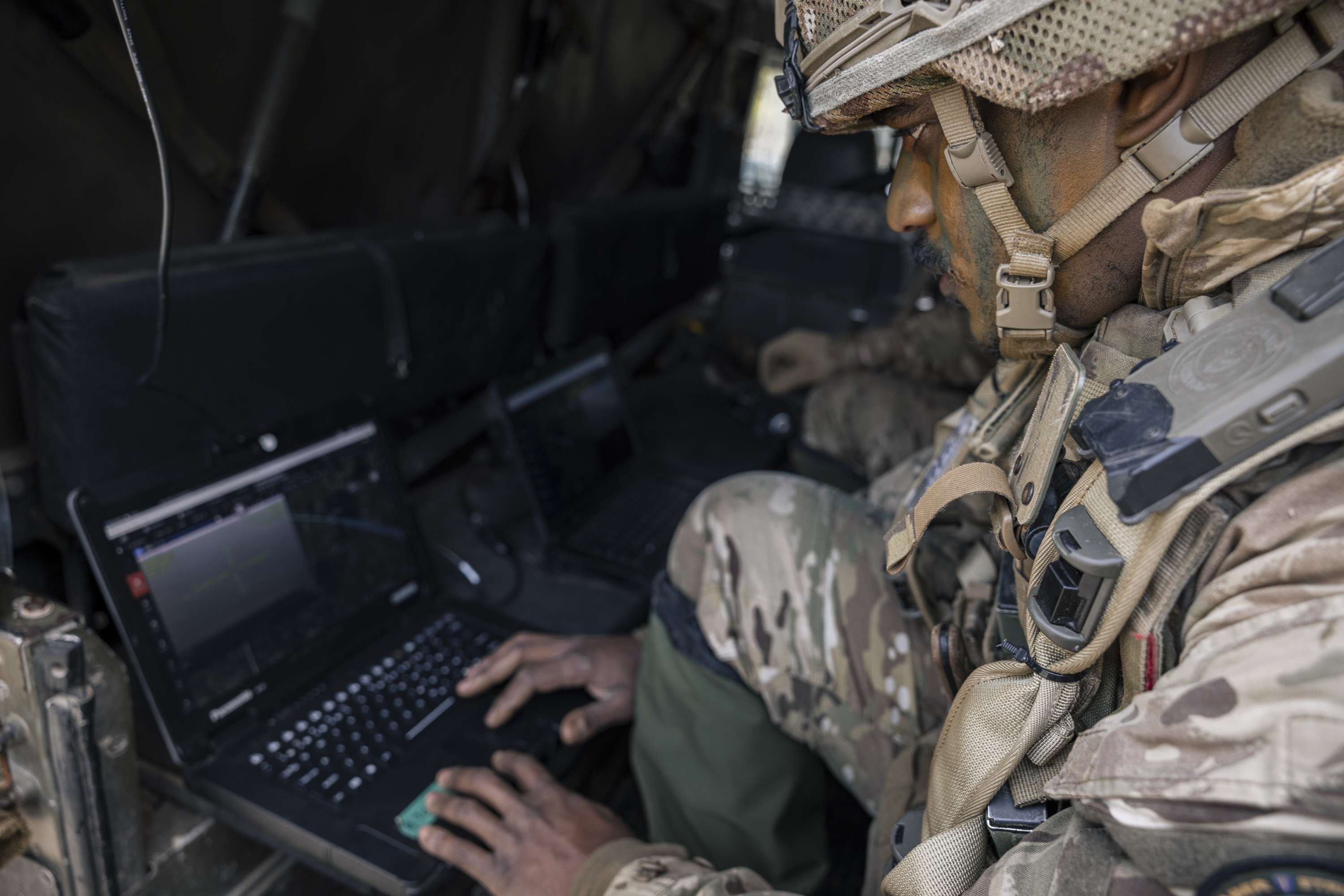 A person in military gear is seated inside a vehicle, focused on using a laptop. The setting suggests a high-tech, tactical environment with multiple laptops visible. The person appears to be engaged in a task that requires attention to the laptop screen.
