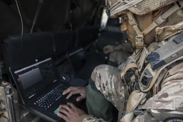 A person in military gear is seated inside a vehicle, focused on using a laptop. The setting suggests a high-tech, tactical environment with multiple laptops visible. The person appears to be engaged in a task that requires attention to the laptop screen.