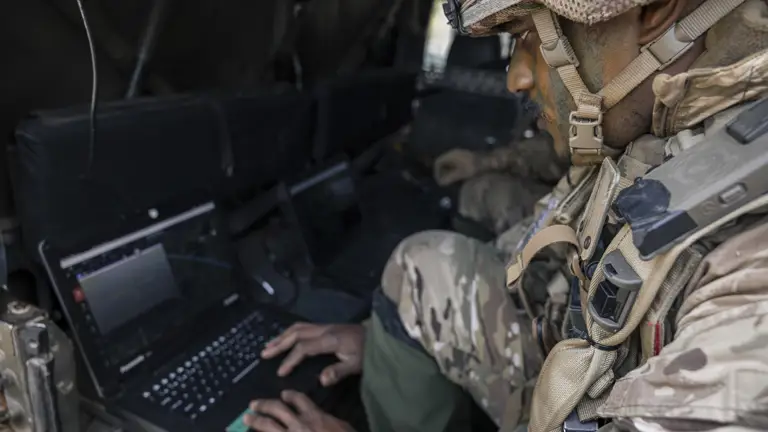 A person in military gear is seated inside a vehicle, focused on using a laptop. The setting suggests a high-tech, tactical environment with multiple laptops visible. The person appears to be engaged in a task that requires attention to the laptop screen.