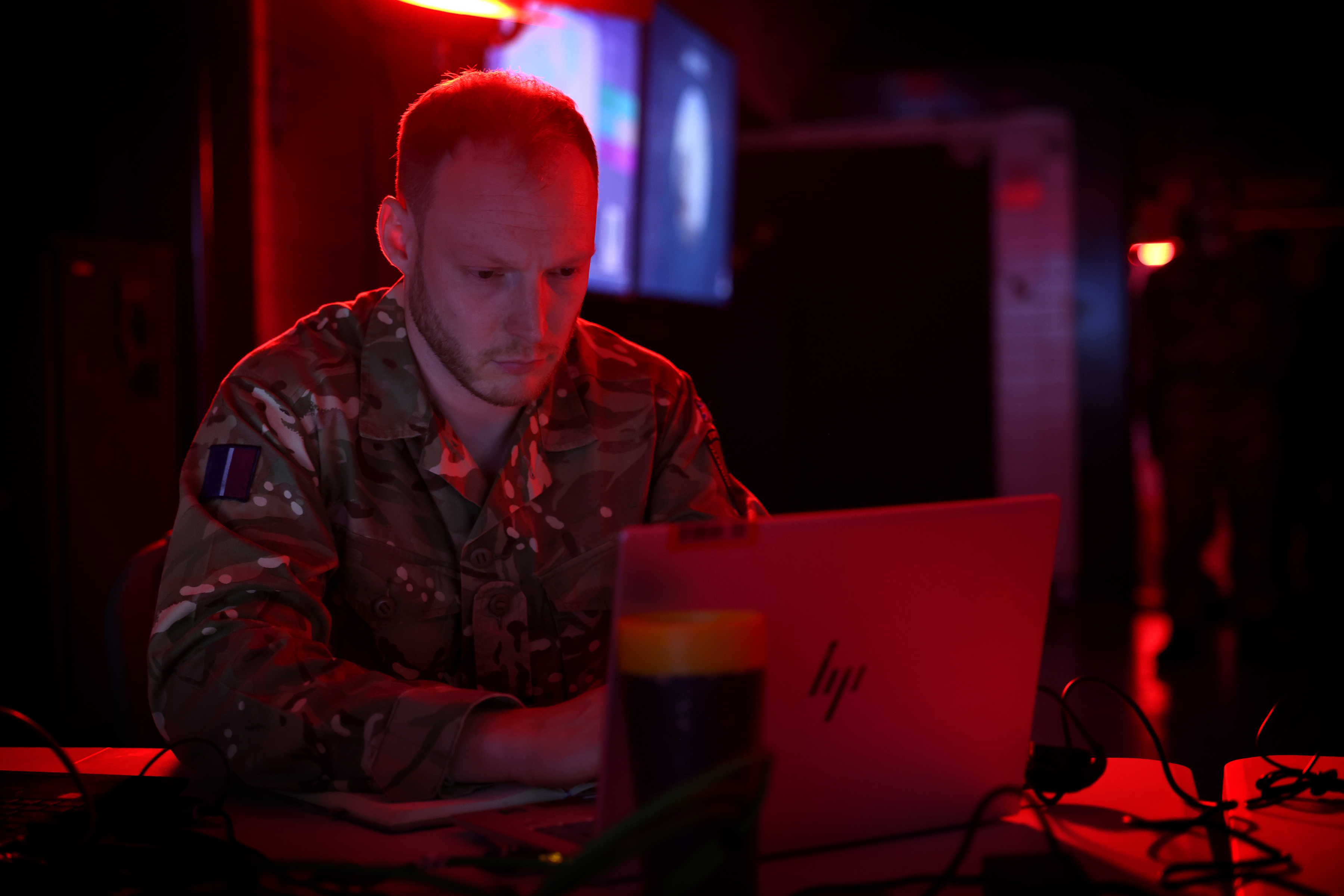 Person in military camouflage using a HP laptop in a dimly lit room with red lighting.