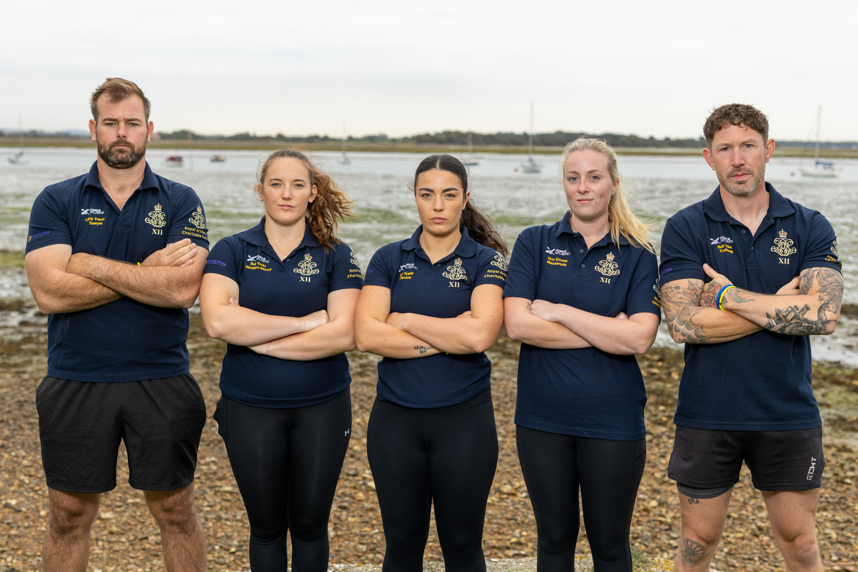 A team of five Army soldiers, two men and three women, are standing in a line wearing dark blue shirts with an Army cap badge on it. They are standing on a shoreline with an estuary in the background.