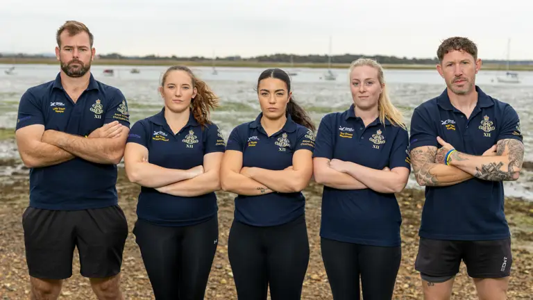 A team of five Army soldiers, two men and three women, are standing in a line wearing dark blue shirts with an Army cap badge on it. They are standing on a shoreline with an estuary in the background.