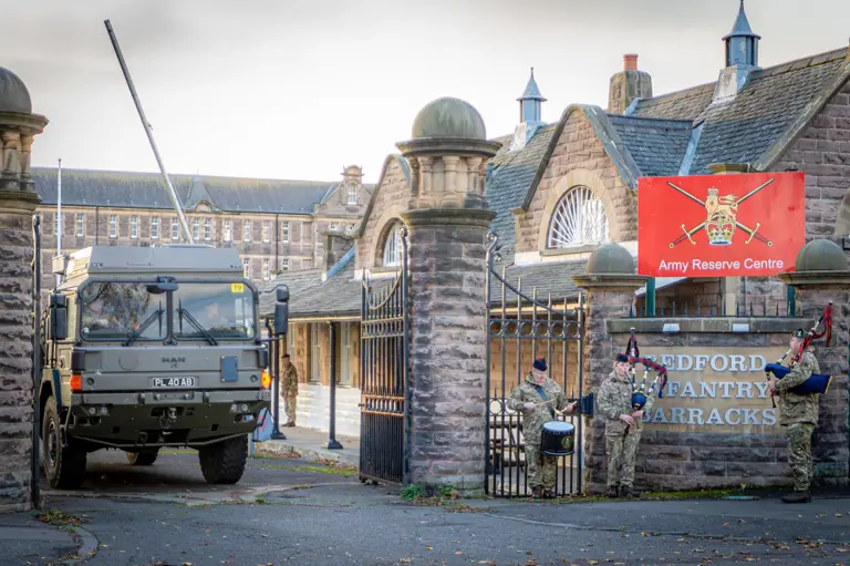 Military truck near stone gate of Bedford Infantry Barracks. Soldiers in uniform play bagpipes and drum, creating a ceremonial atmosphere.