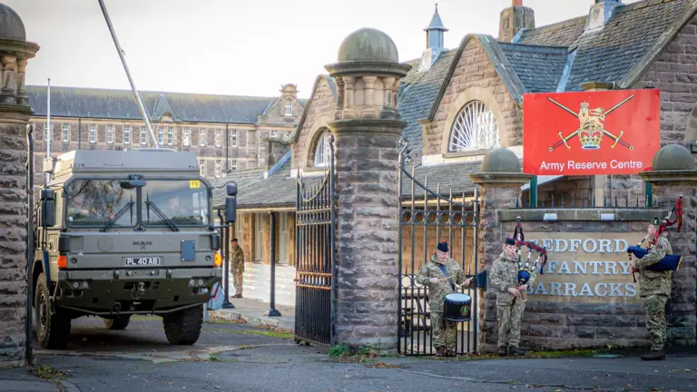 Military truck near stone gate of Bedford Infantry Barracks. Soldiers in uniform play bagpipes and drum, creating a ceremonial atmosphere.