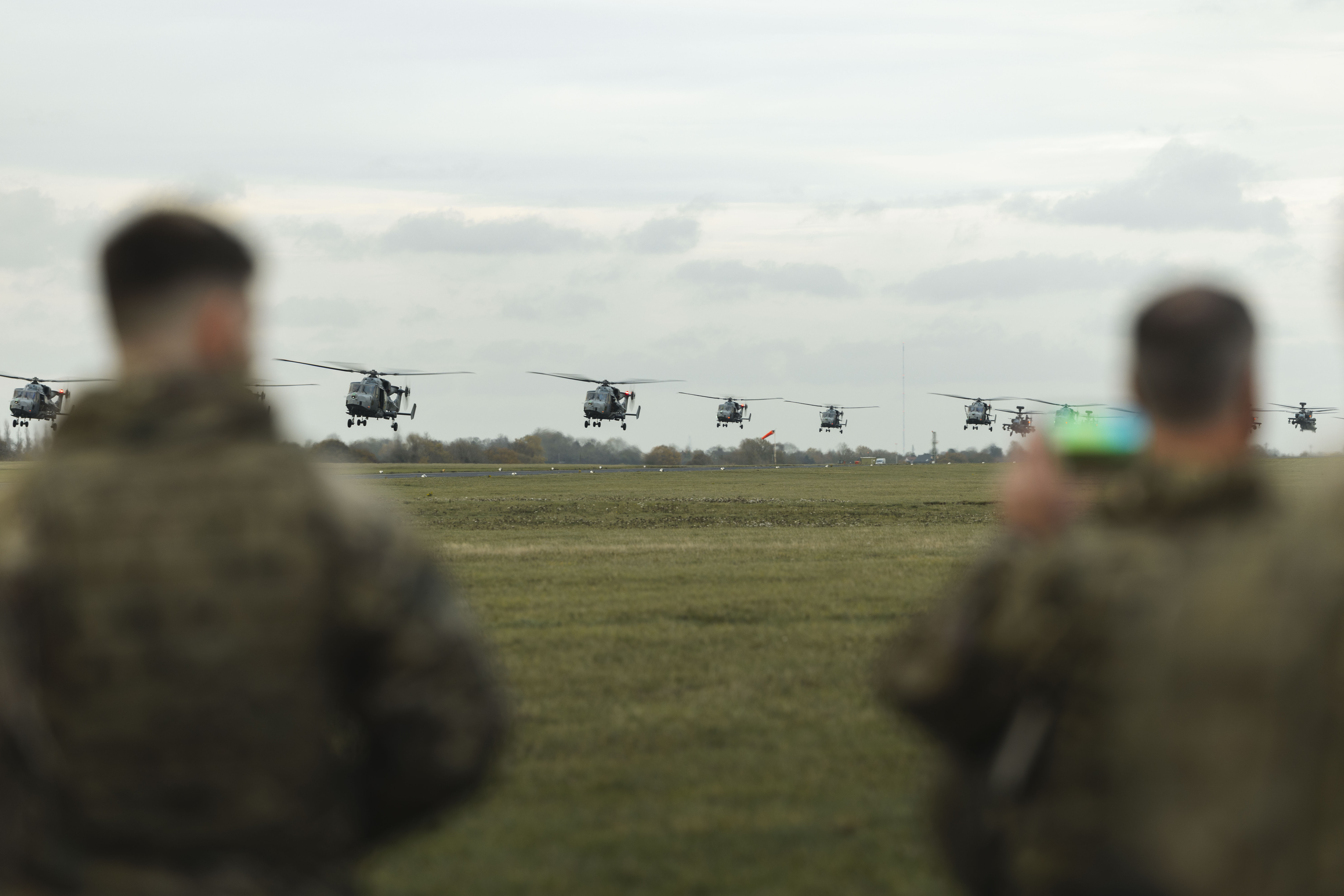 Eight military helicopters flying low in formation over an open field with two soldiers observing in the foreground.