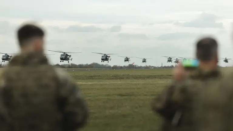 Eight military helicopters flying low in formation over an open field with two soldiers observing in the foreground.