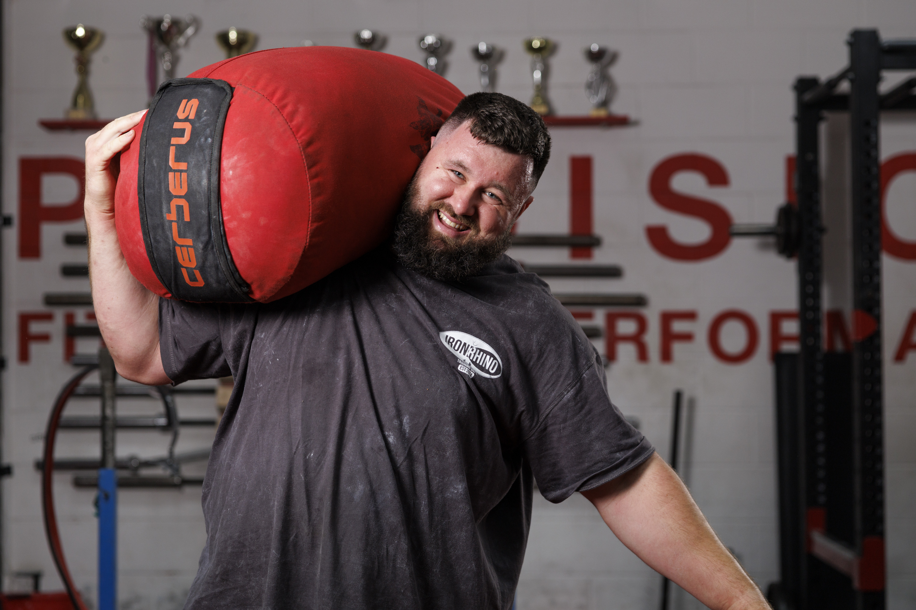 A person is smiling and holding a large, red Cerberus sandbag over their shoulder. They are standing in a gym with weightlifting equipment and several trophies on a shelf in the background.