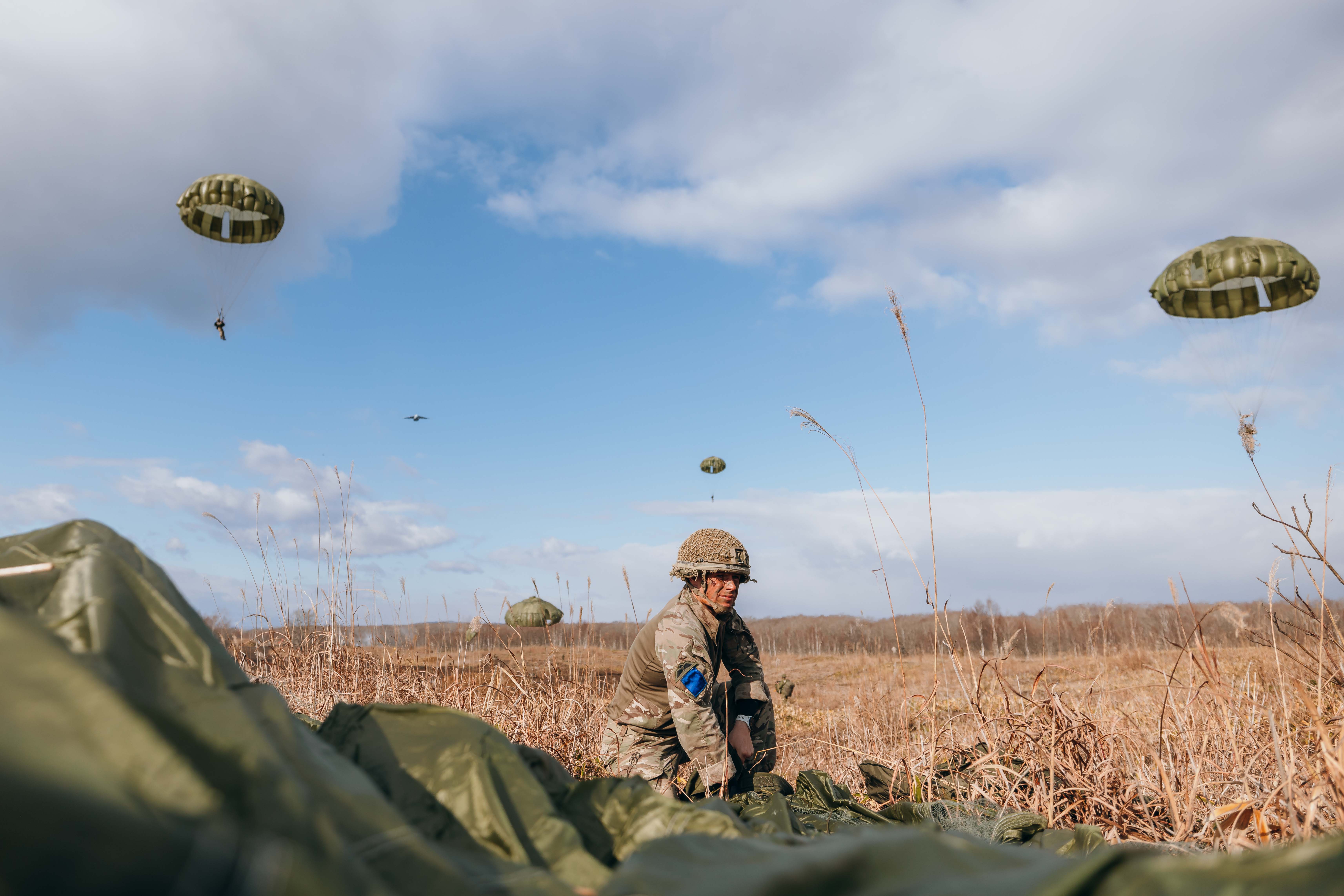 Soldier in camouflage gear kneeling in a dry grassy field with multiple parachutes descending in the blue sky above.