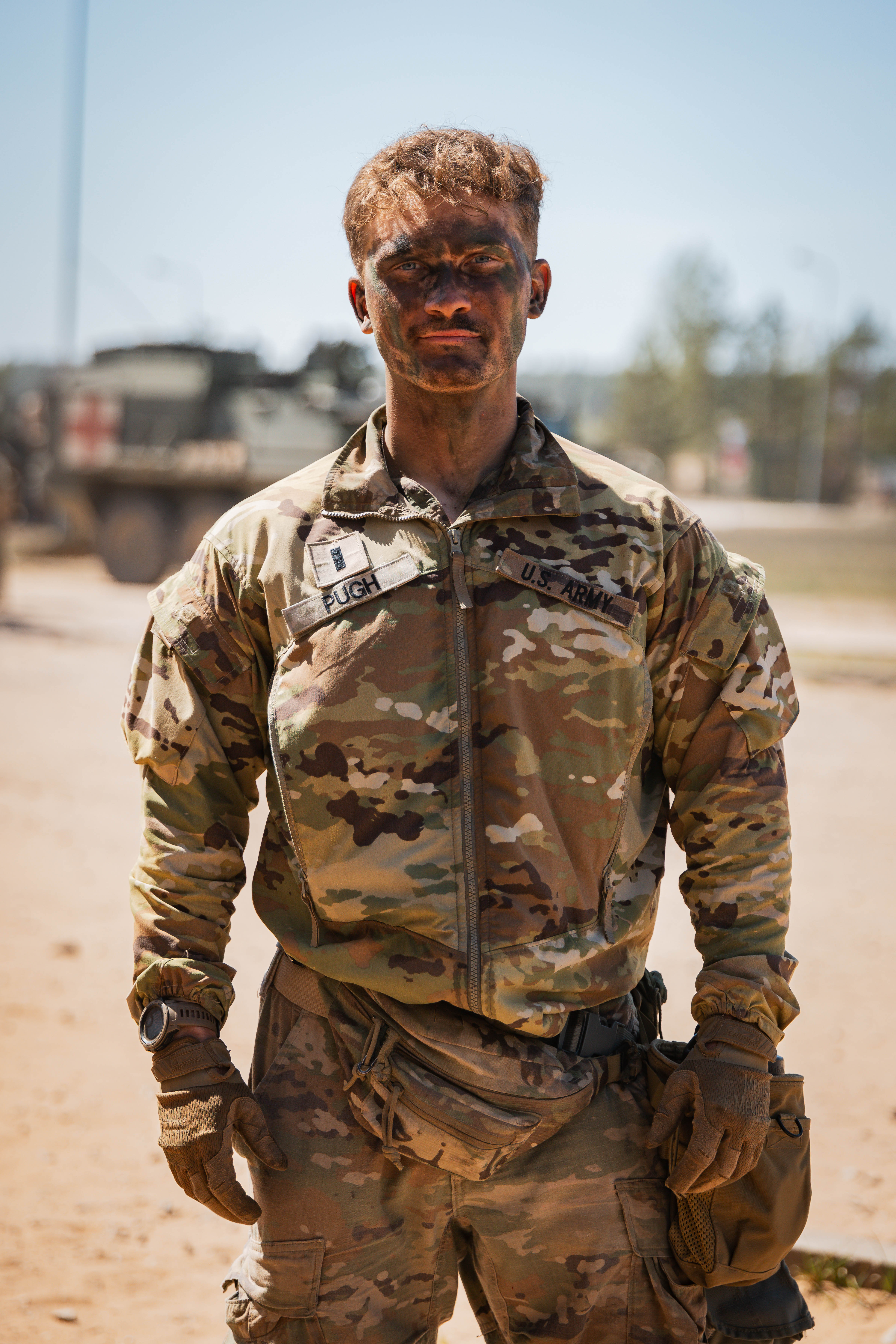 A soldier in camouflage uniform stands outdoors, face covered in dirt. Sunlight accentuates determination. Blurred military vehicles in the background.
