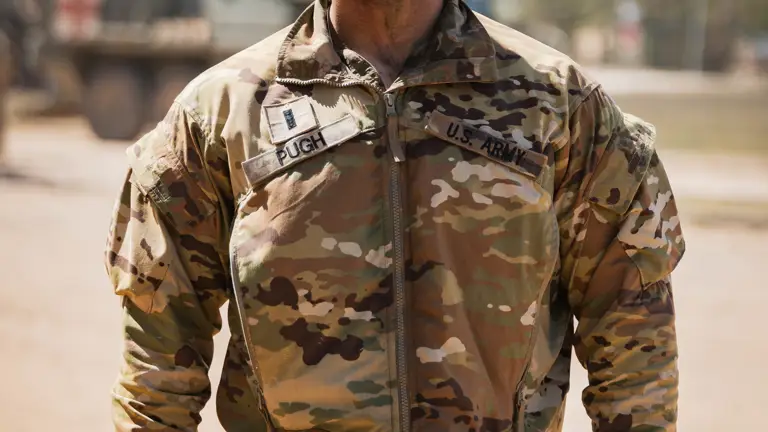 A soldier in camouflage uniform stands outdoors, face covered in dirt. Sunlight accentuates determination. Blurred military vehicles in the background.