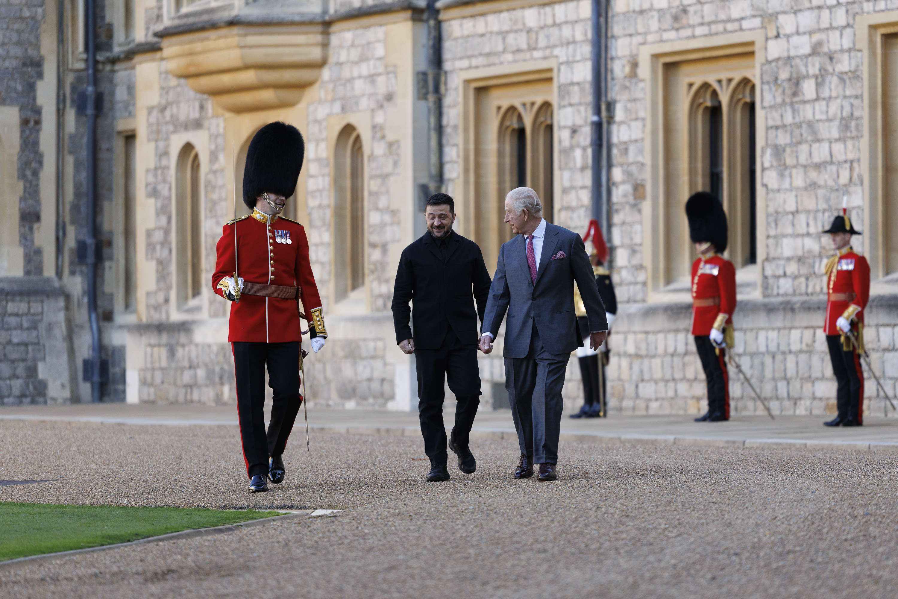 Two men, one in a suit and the other in a black coat, walk together outside a historic stone building.