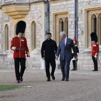 Two men, one in a suit and the other in a black coat, walk together outside a historic stone building.
